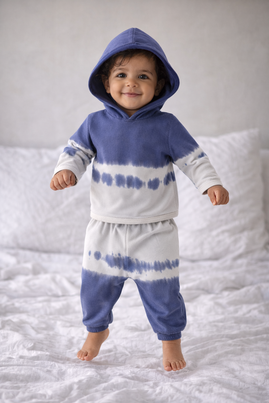 Child wearing a blue and white tie-dye hoodie and pants on a white bed.
