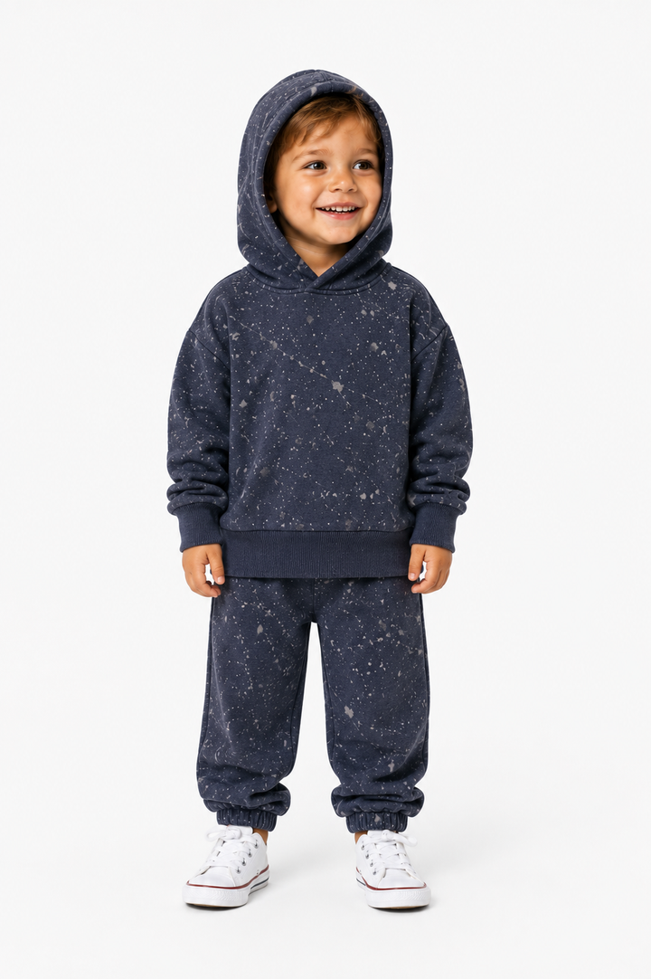 A young child wearing a matching navy blue hooded sweatshirt and jogger pants set with a white speckled pattern, paired with white sneakers, standing against a plain white background.