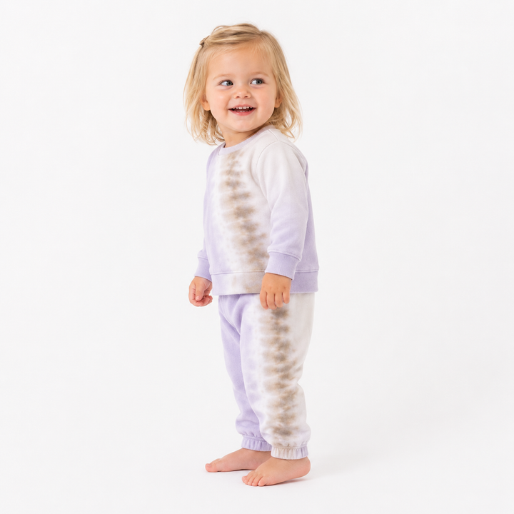 A young toddler girl with blonde hair stands barefoot against a plain white studio background, wearing a matching two-piece pajama set with a light lavender and white tie-dye pattern.
