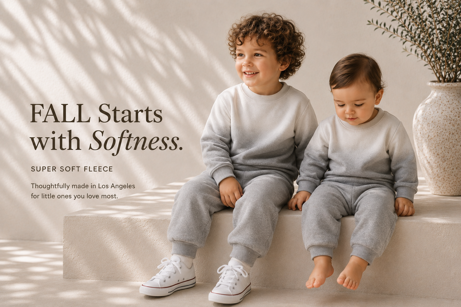 Two children wearing matching light gray fleece sweatshirt and jogger pants sets sit on a beige stone bench in a softly lit room. Text on the left reads, 'FALL Starts with Softness.