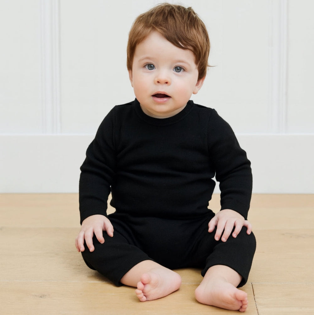 Baby wearing a black outfit sitting on a wooden floor with a white wall background