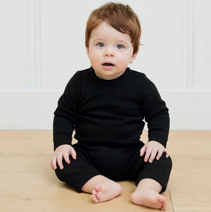 Baby wearing a black outfit sitting on a wooden floor with a white wall background