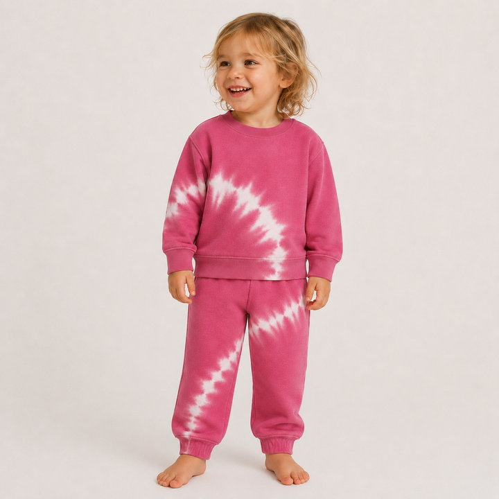 A baby wearing a matching pink tie-dye sweatshirt and jogger pants set, standing against a plain light gray studio background.