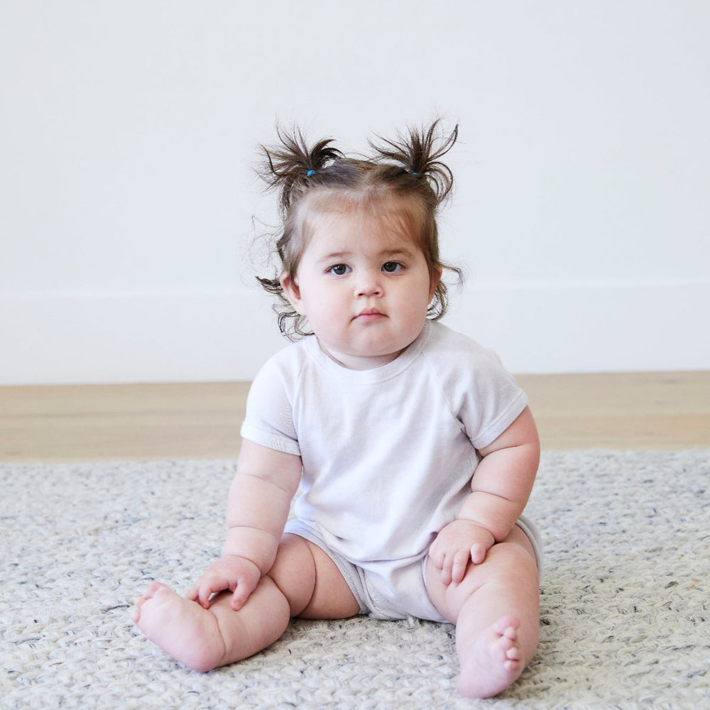 Baby sitting on a carpeted floor wearing a plain white t-shirt and light-colored pants.