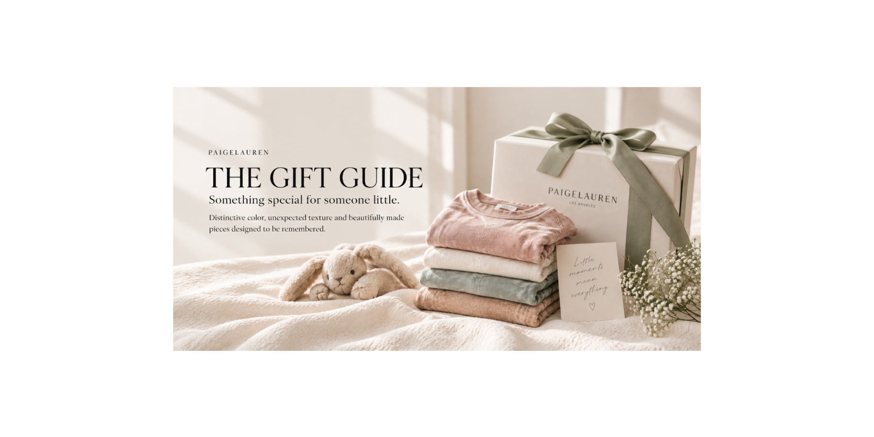A styled gift set from PaigeLauren on a soft cream bed, featuring a white gift box with a sage green ribbon, a stack of folded pastel garments in pink, cream, and sage green, a plush bunny toy, and a handwritten note. Text on the left reads 'PAIGE LAUREN THE GIFT GUIDE'.