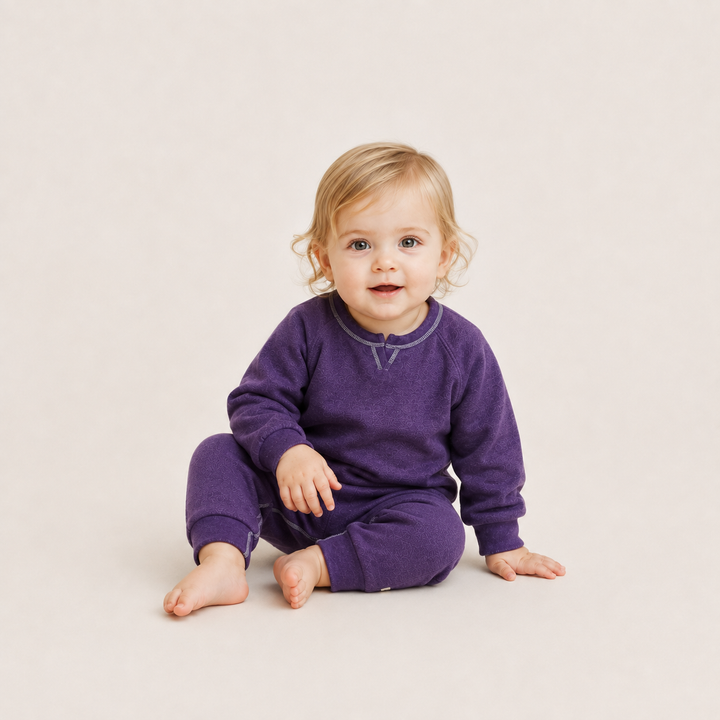 A toddler with blonde hair sits cross-legged on a light beige studio background, wearing a matching purple long-sleeve top and pants set with a subtle contrasting collar detail.