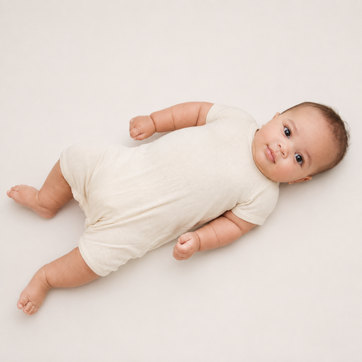 Baby lying down wearing the PAIGELAUREN Garment Dye Ribbed Shortall Onesie. Beige