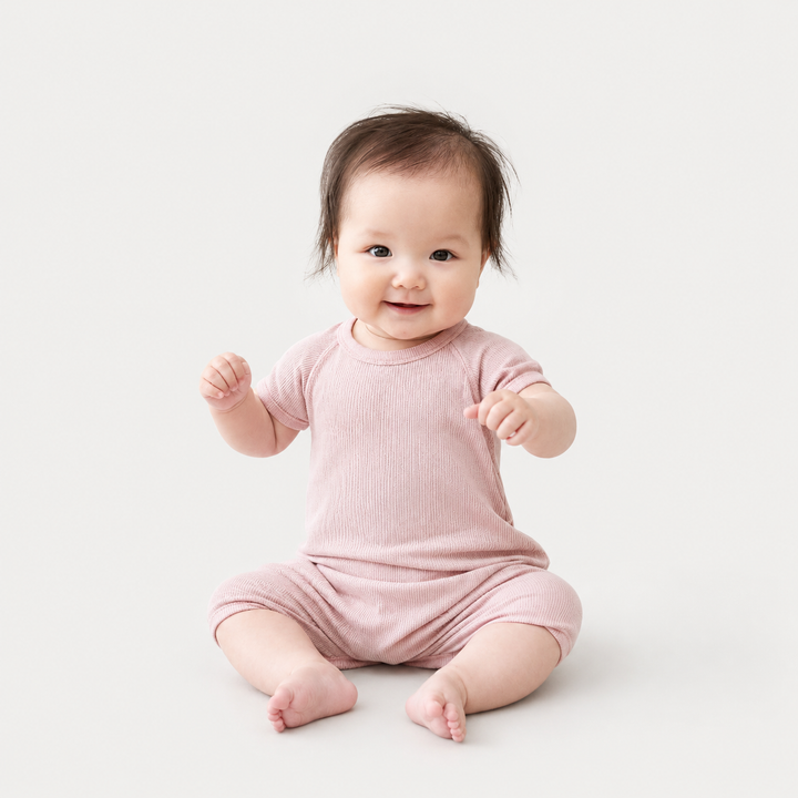 Baby wearing the PAIGELAUREN Garment Dye Ribbed Shortall Onesie in Blush Pink while sitting