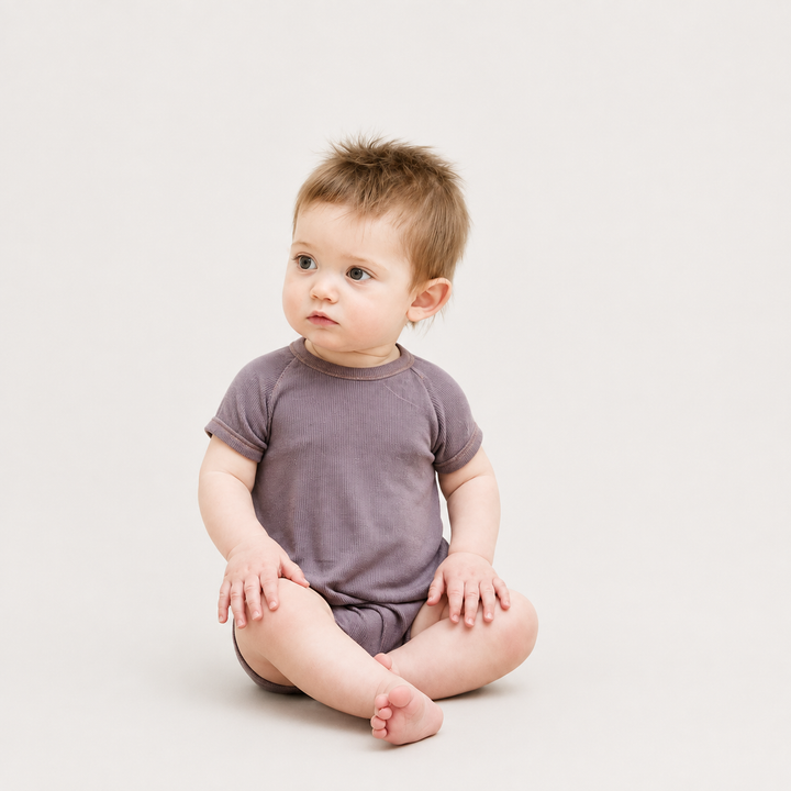 Baby wearing the PAIGELAUREN Garment Dye Ribbed Shortall Onesie in Plum while sitting