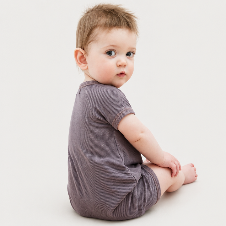 Back view of a baby wearing the PAIGELAUREN Garment Dye Ribbed Shortall Onesie in Plum