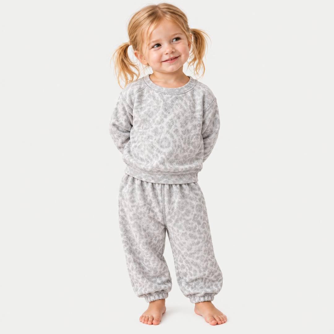A toddler child with blonde pigtails stands barefoot against a light gray background, wearing a matching light gray and white animal-print sweatshirt and sweatpants set.