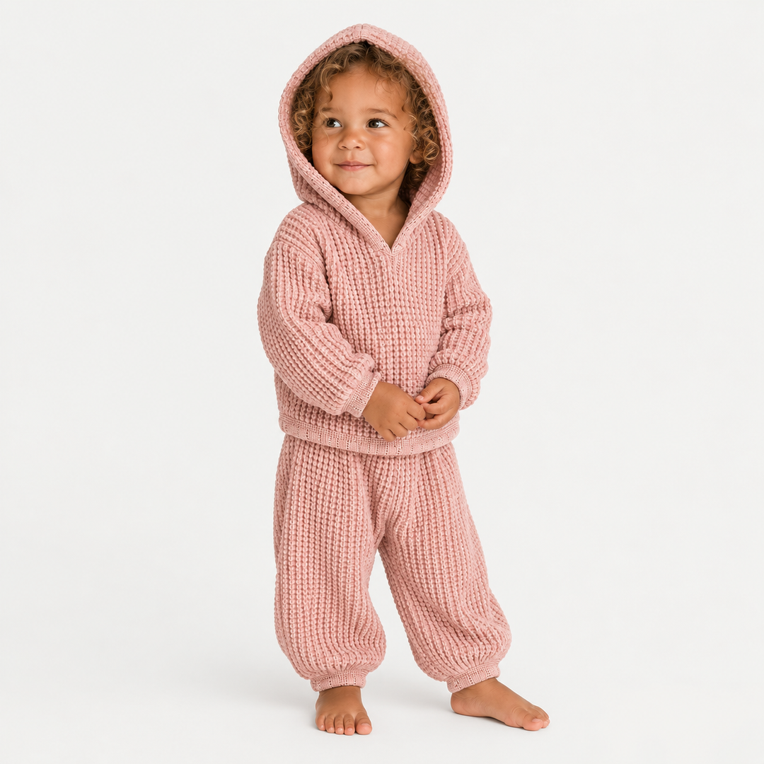 A toddler with curly hair stands barefoot wearing a dusty pink waffle-knit hooded sweatshirt and matching pants against a clean white background.