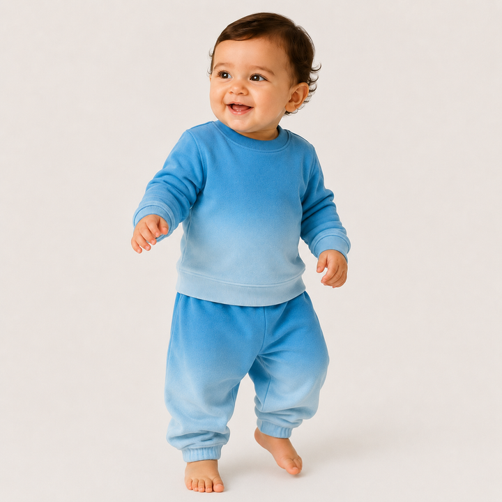 A smiling baby wearing a matching two-piece blue gradient sweatshirt and sweatpants set, standing barefoot against a plain light background.