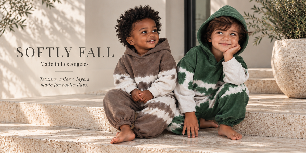 Two toddlers wearing PAIGELAUREN cocoa and hunter green hand-dyed loungewear sets, seated on travertine steps in a softly lit California setting.