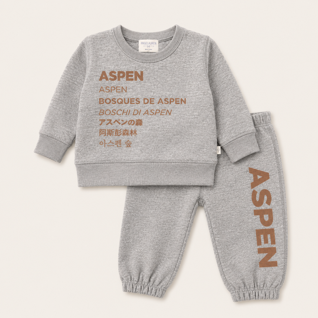 A two-piece kids' loungewear set by Aspen in heather gray, featuring a sweatshirt and jogger pants with brown text graphics. The sweatshirt features 'ASPEN BOSQUES DE ASPEN BOSCHI DI ASPEN' along with Japanese and Korean text.