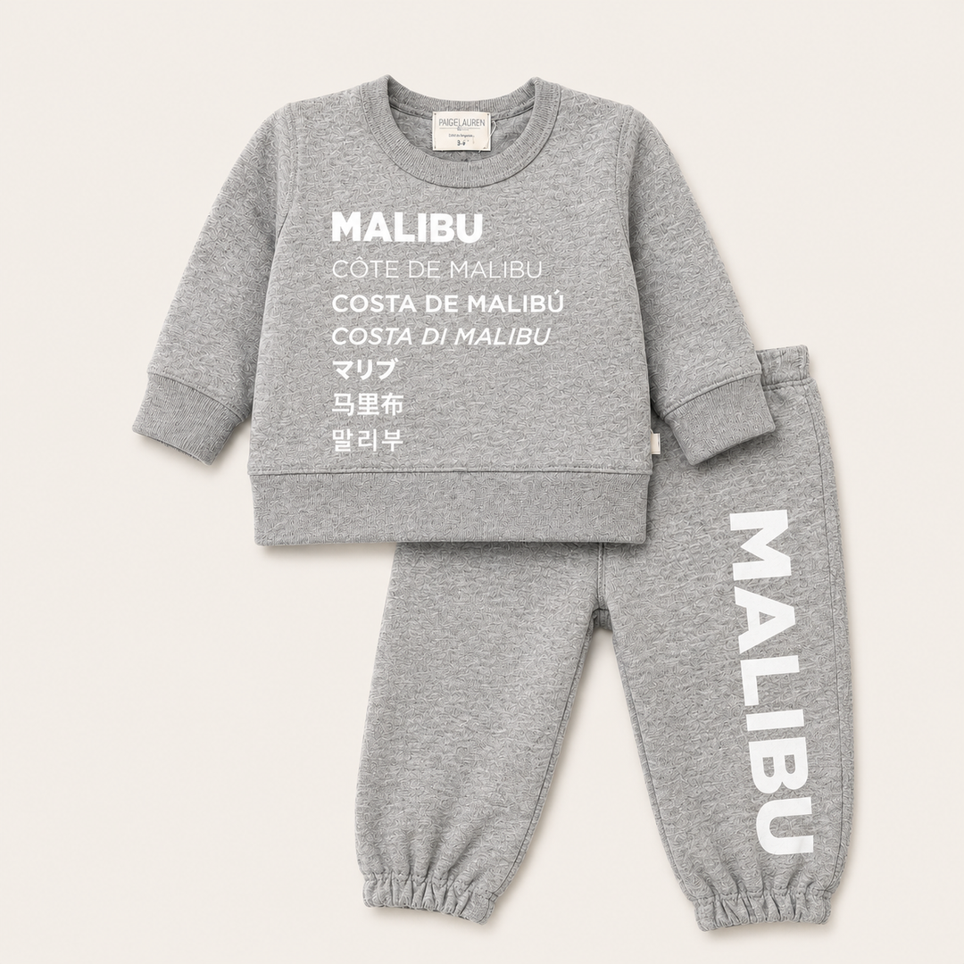 A two-piece kids' loungewear set in heather gray, featuring a long-sleeve sweatshirt and matching jogger pants. The sweatshirt has 'MALIBU' printed in white across the chest with translations in multiple languages below.