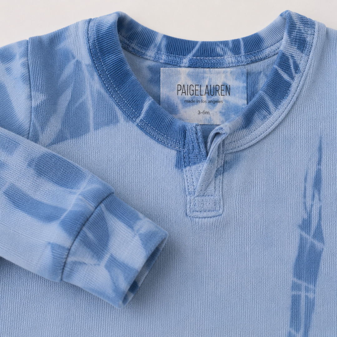 A close-up of a light blue tie-dye long-sleeve henley shirt by PaigeLauren, showing the collar label and folded sleeve details.