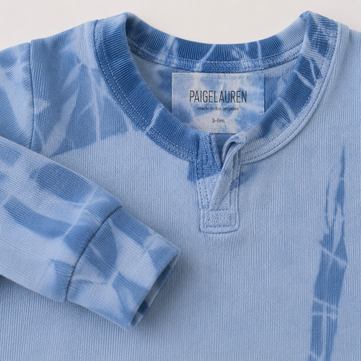 A close-up of a light blue tie-dye long-sleeve henley shirt by PaigeLauren, showing the collar label and folded sleeve details.